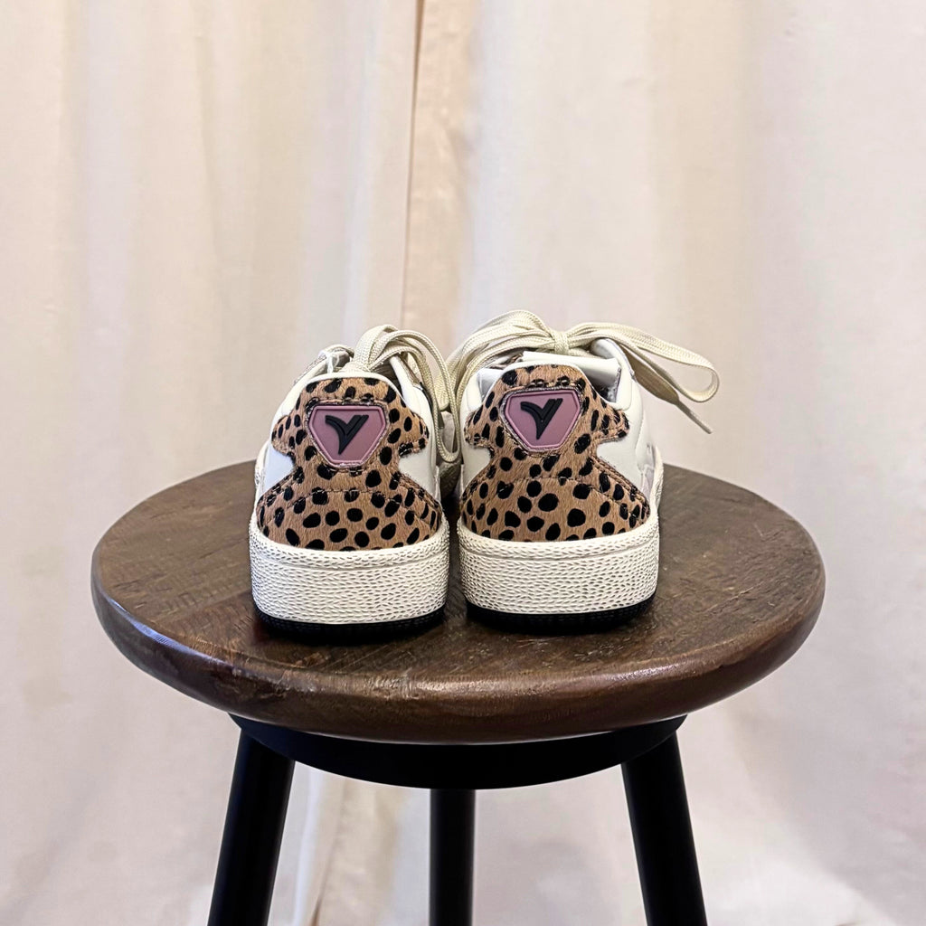 Pair of leopard print sneakers with pink hearts on a wooden stool against a white curtain background