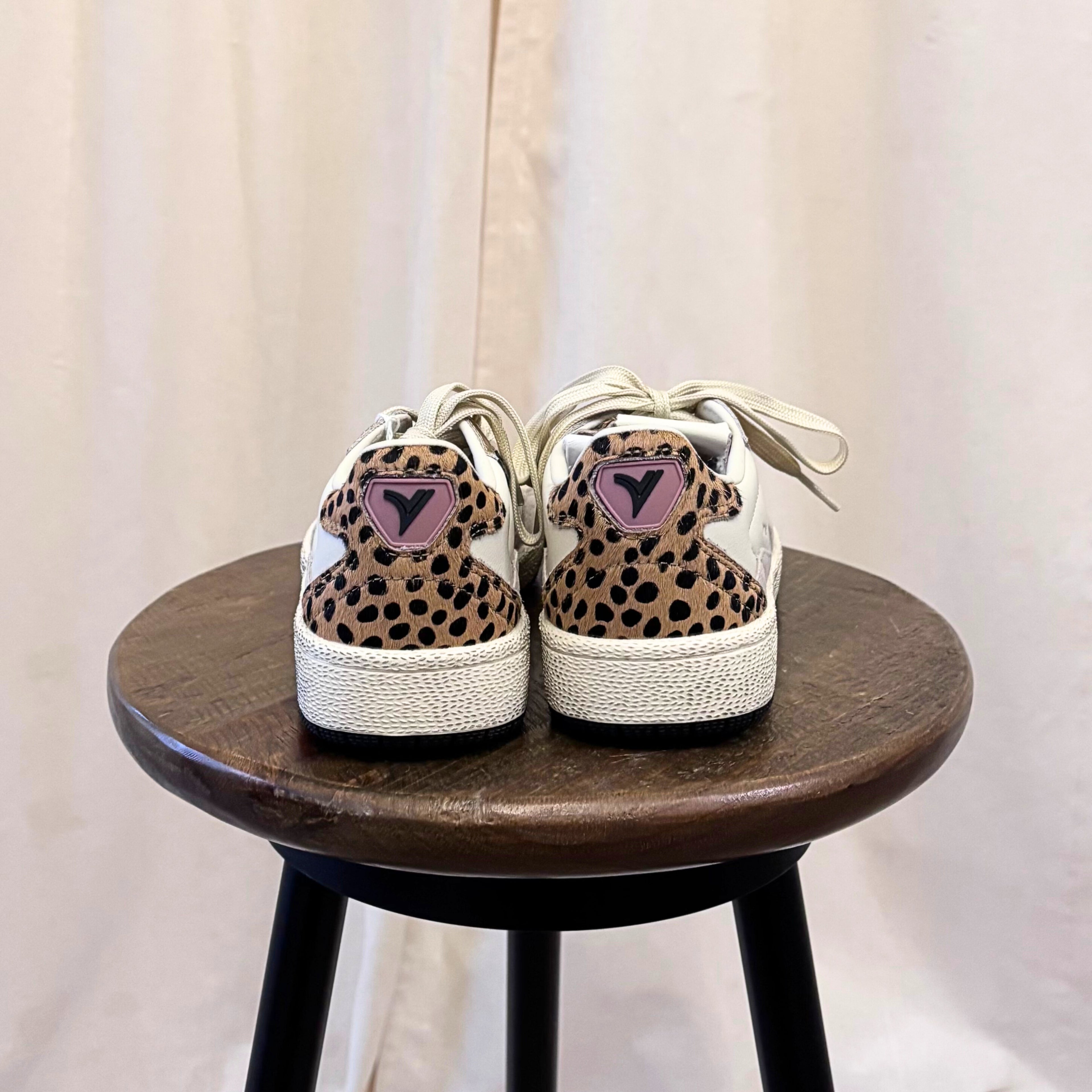 Pair of leopard print sneakers with pink hearts on a wooden stool against a white curtain background