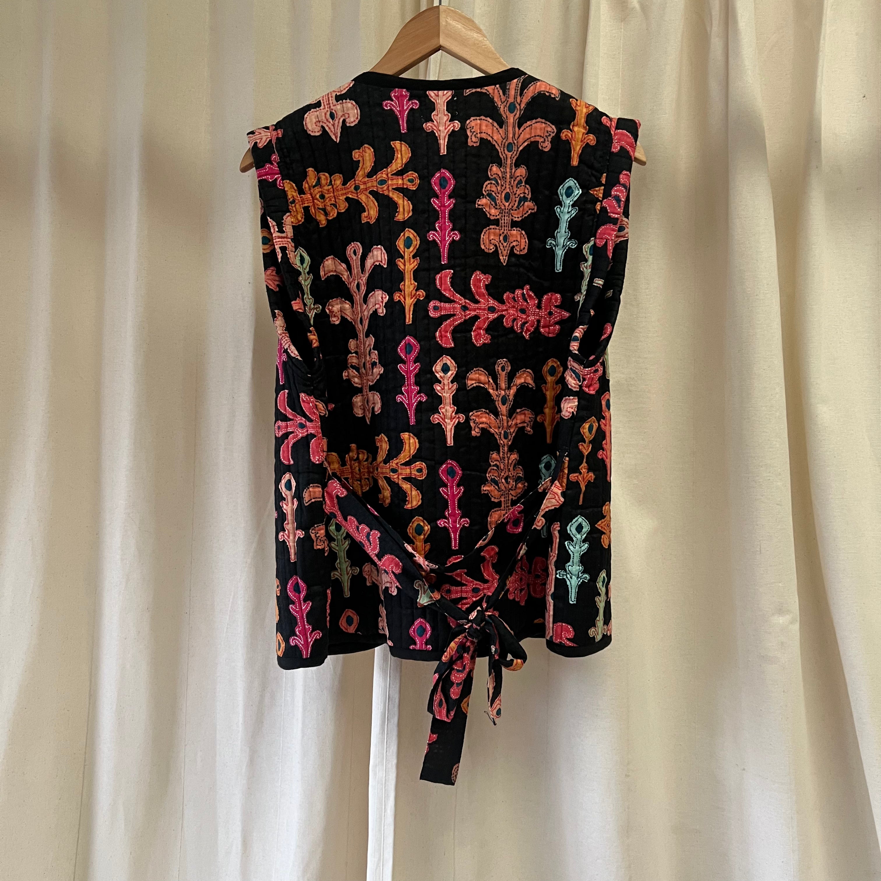 Black sleeveless top with colorful patterns on a hanger against a white curtain background