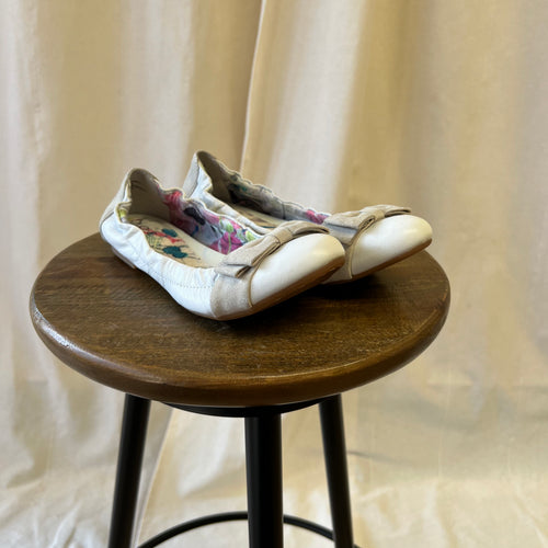 Pair of white flat shoes on a wooden stool against a beige curtain background