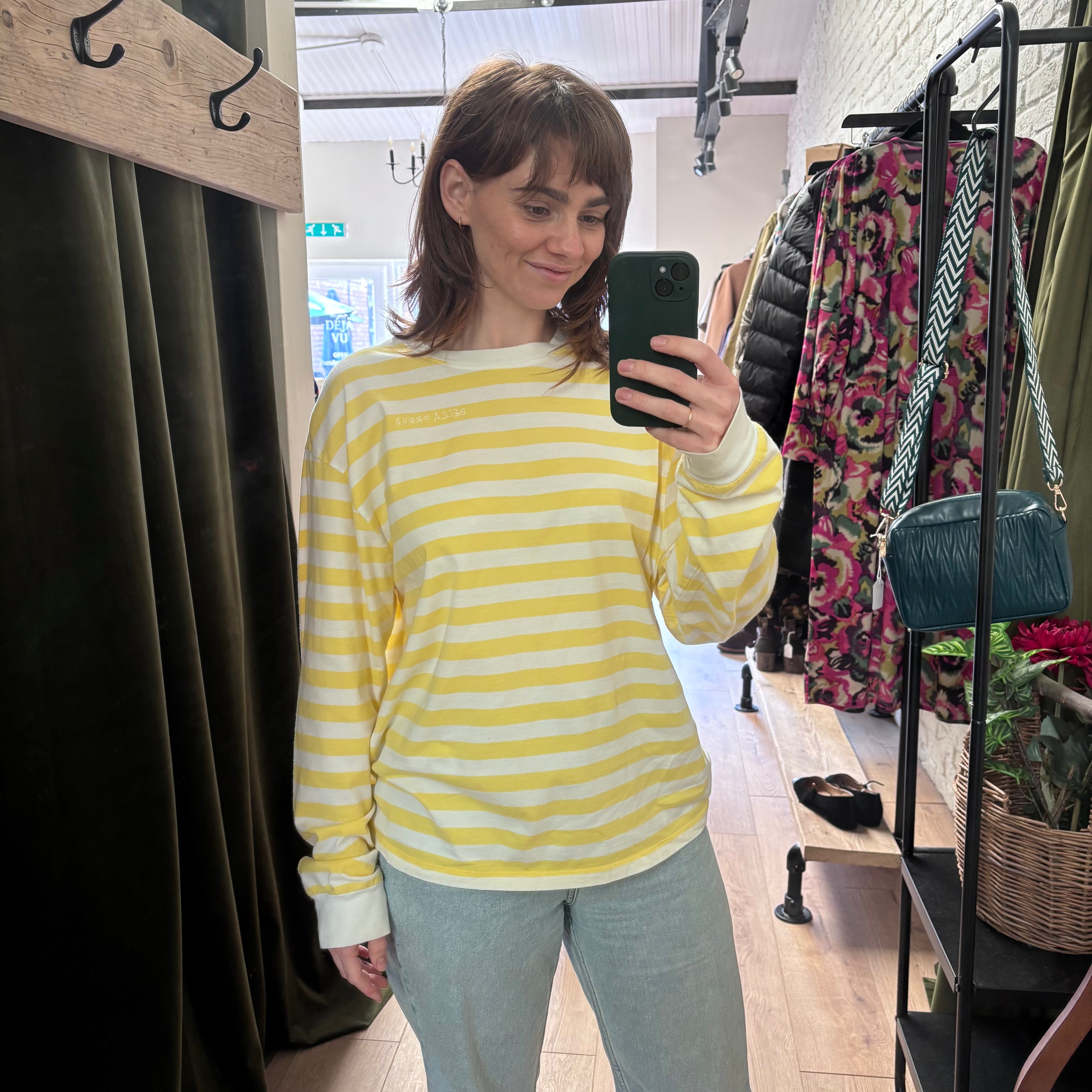 Woman taking a mirror selfie wearing a yellow and white striped sweater in a clothing store.