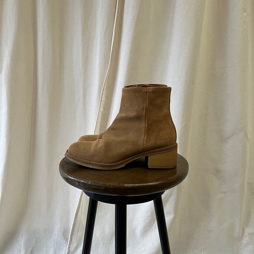 Brown suede ankle boot on a small wooden stool against a white curtain background