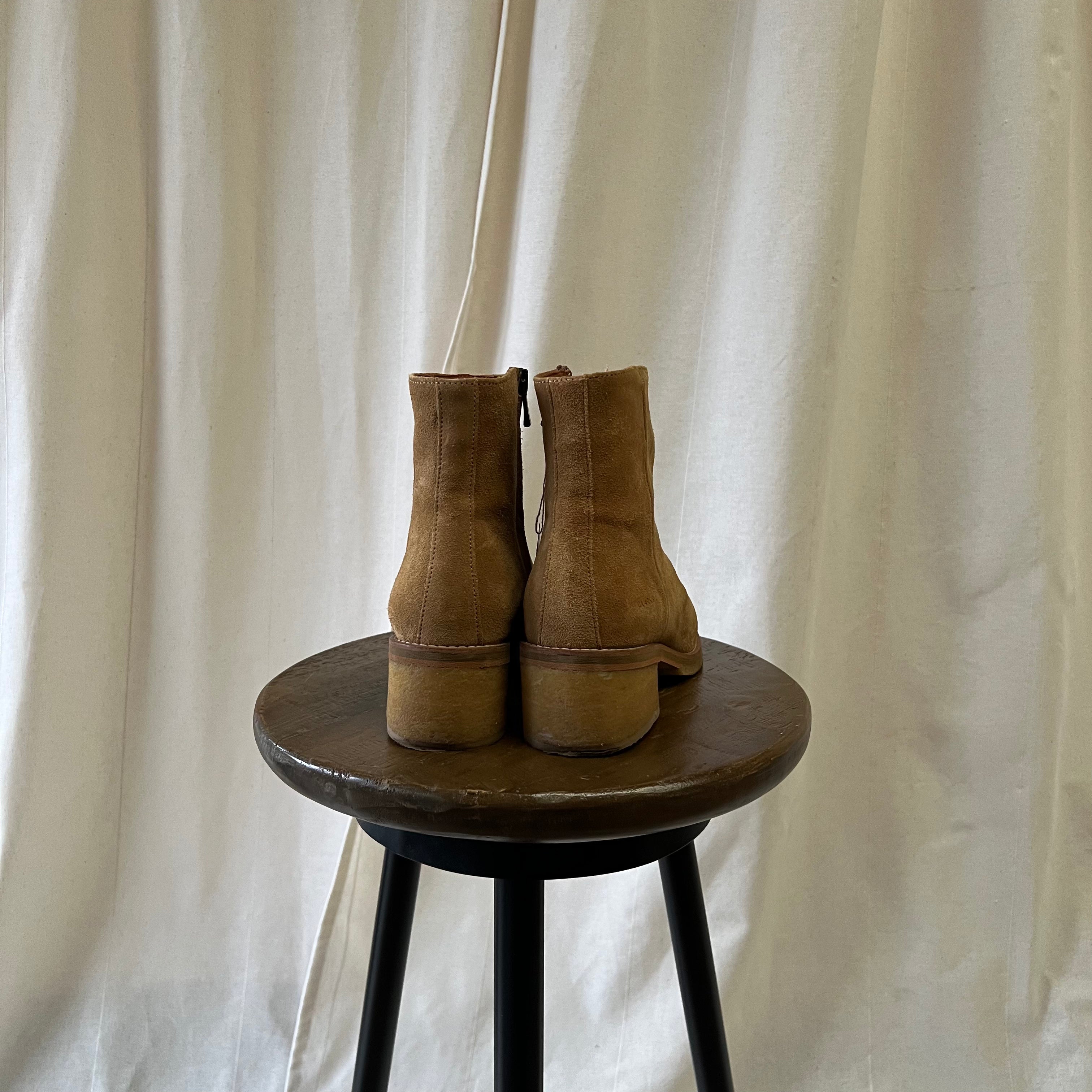 Pair of brown suede boots on a wooden stool against a white curtain background