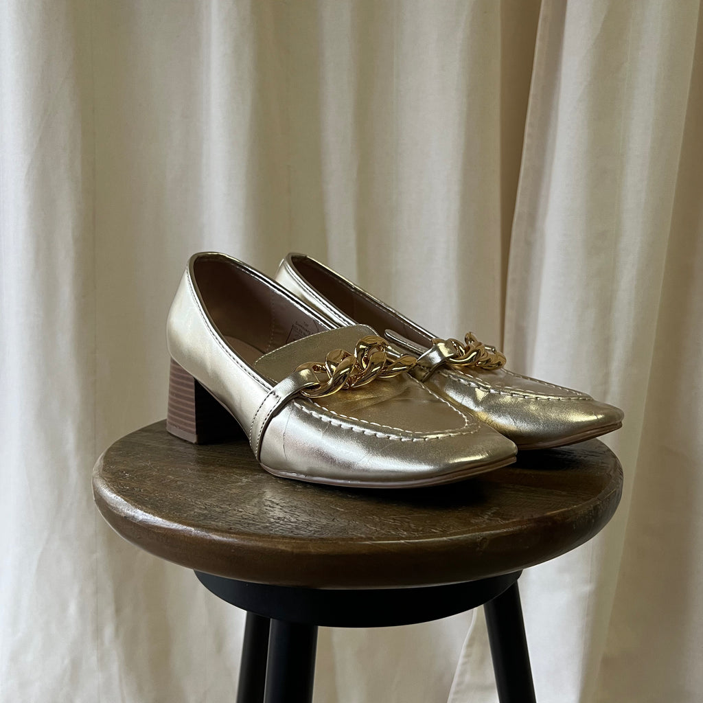 Pair of metallic loafers with gold chain detail on a wooden stool against a neutral curtain background
