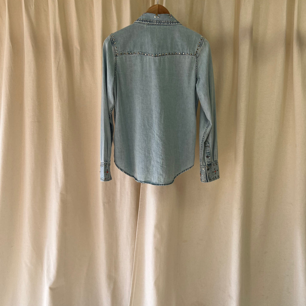 Light blue denim shirt on a wooden hanger against a beige curtain background