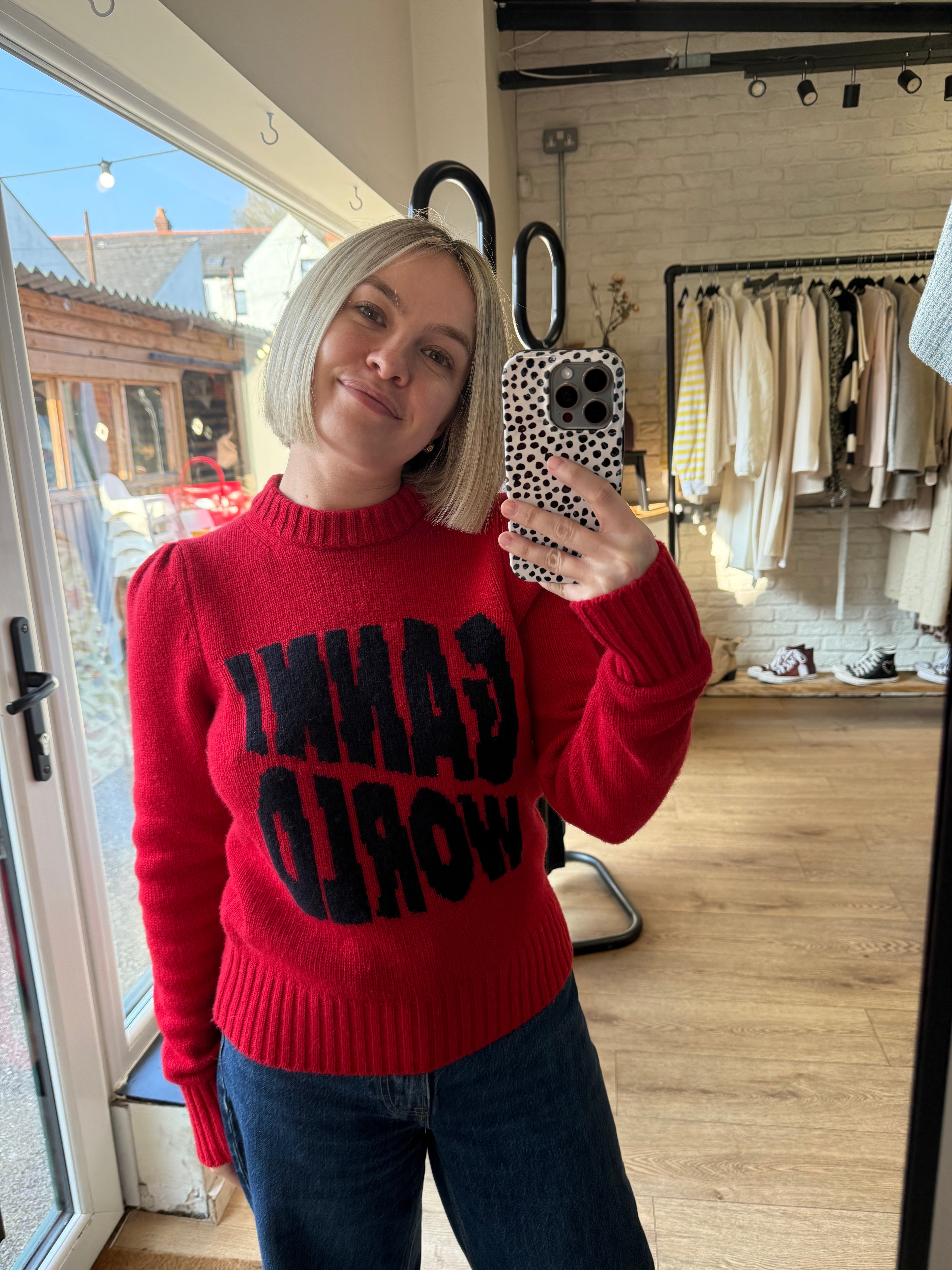 Person wearing a red sweater with text, taking a mirror selfie in a room with clothing racks.