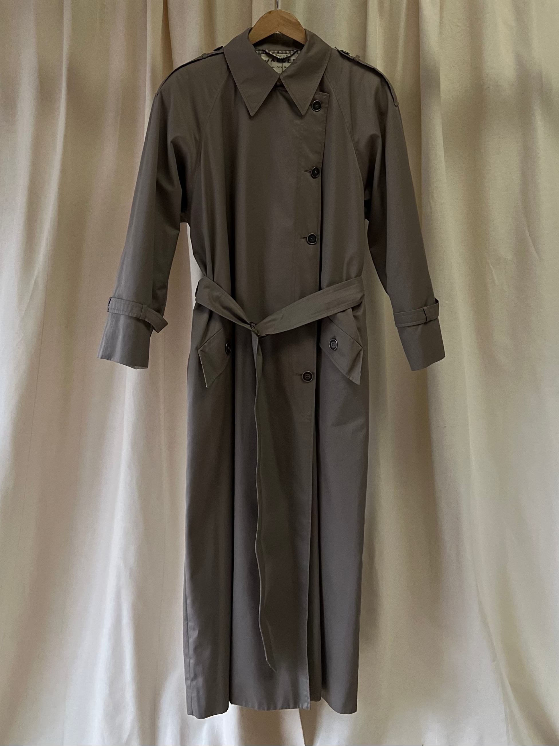 Grey trench coat hanging on a hanger against a white curtain backdrop
