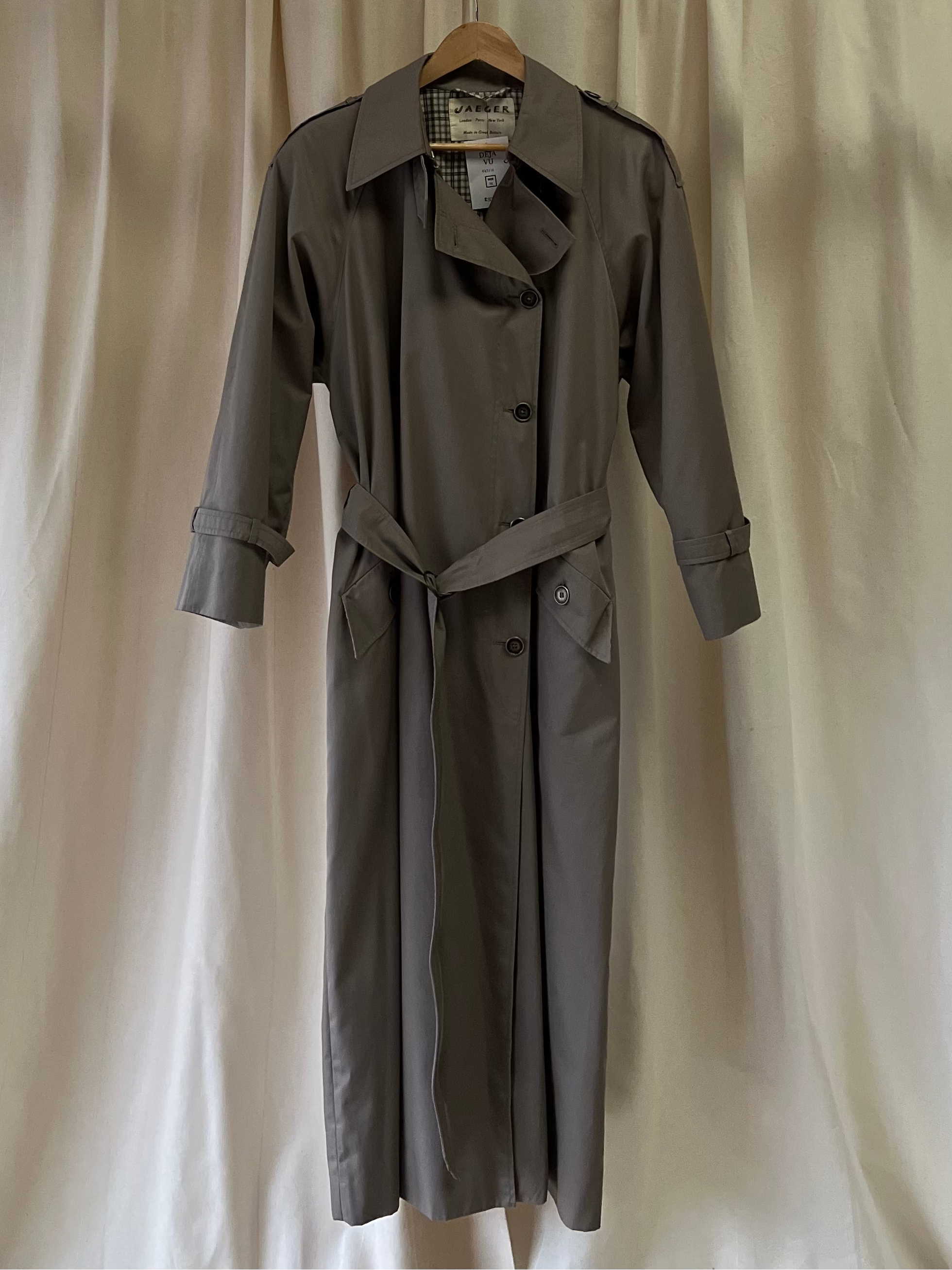 Grey trench coat hanging on a hanger against a white curtain background