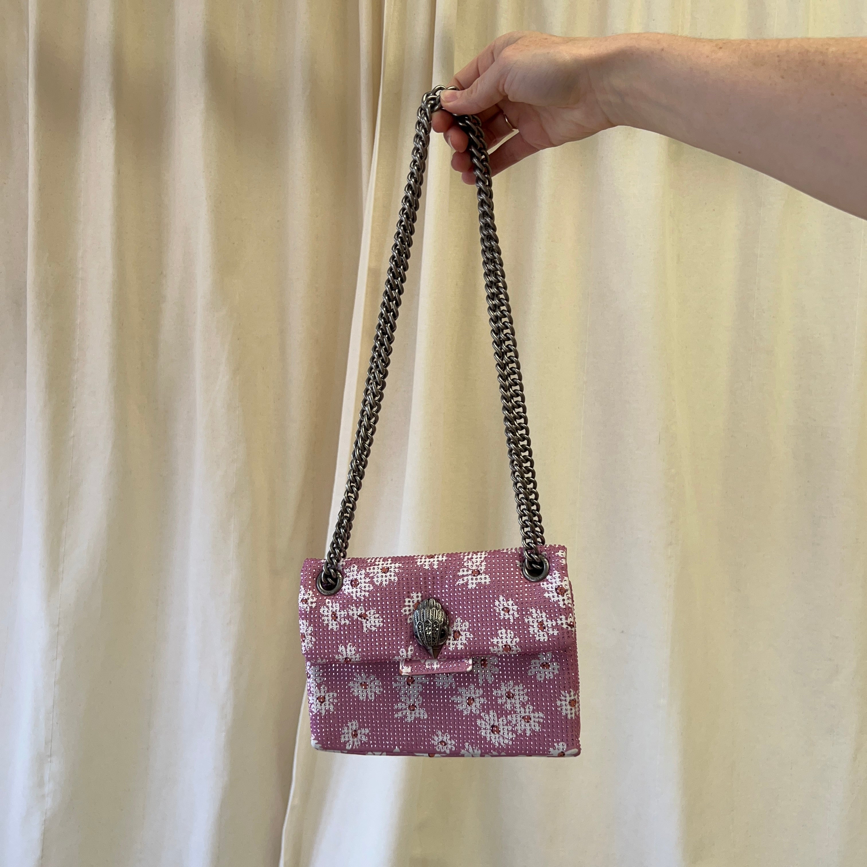 Hand holding a pink floral handbag with a chain strap against a beige curtain background