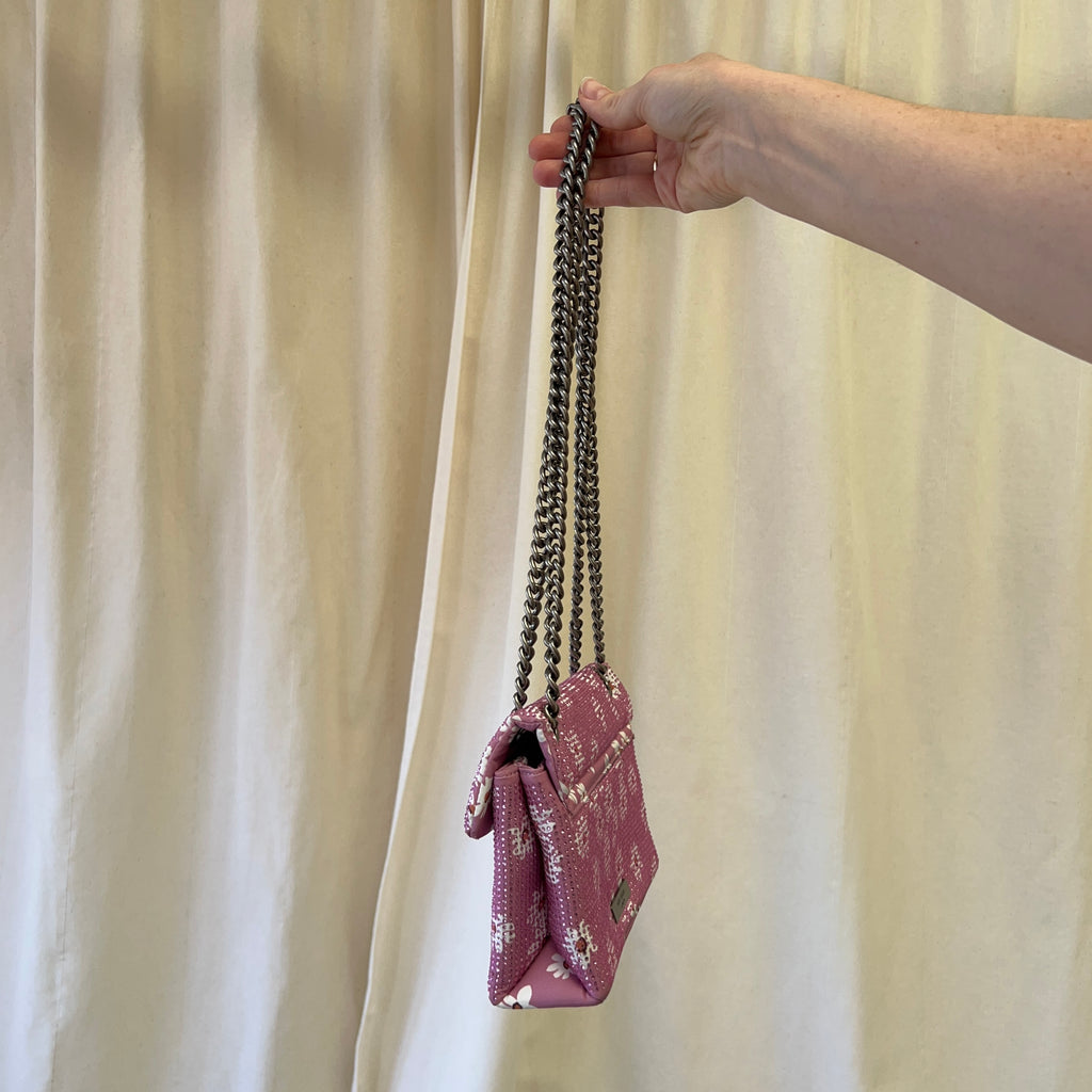 Hand holding a pink floral handbag with a chain strap against a beige curtain background