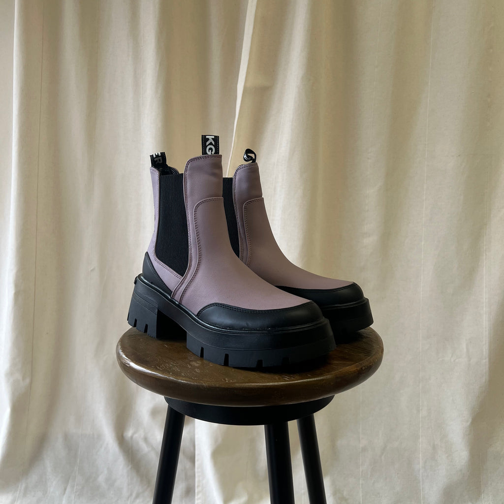 Pair of purple Chelsea boots with black soles on a wooden stool against a beige curtain background