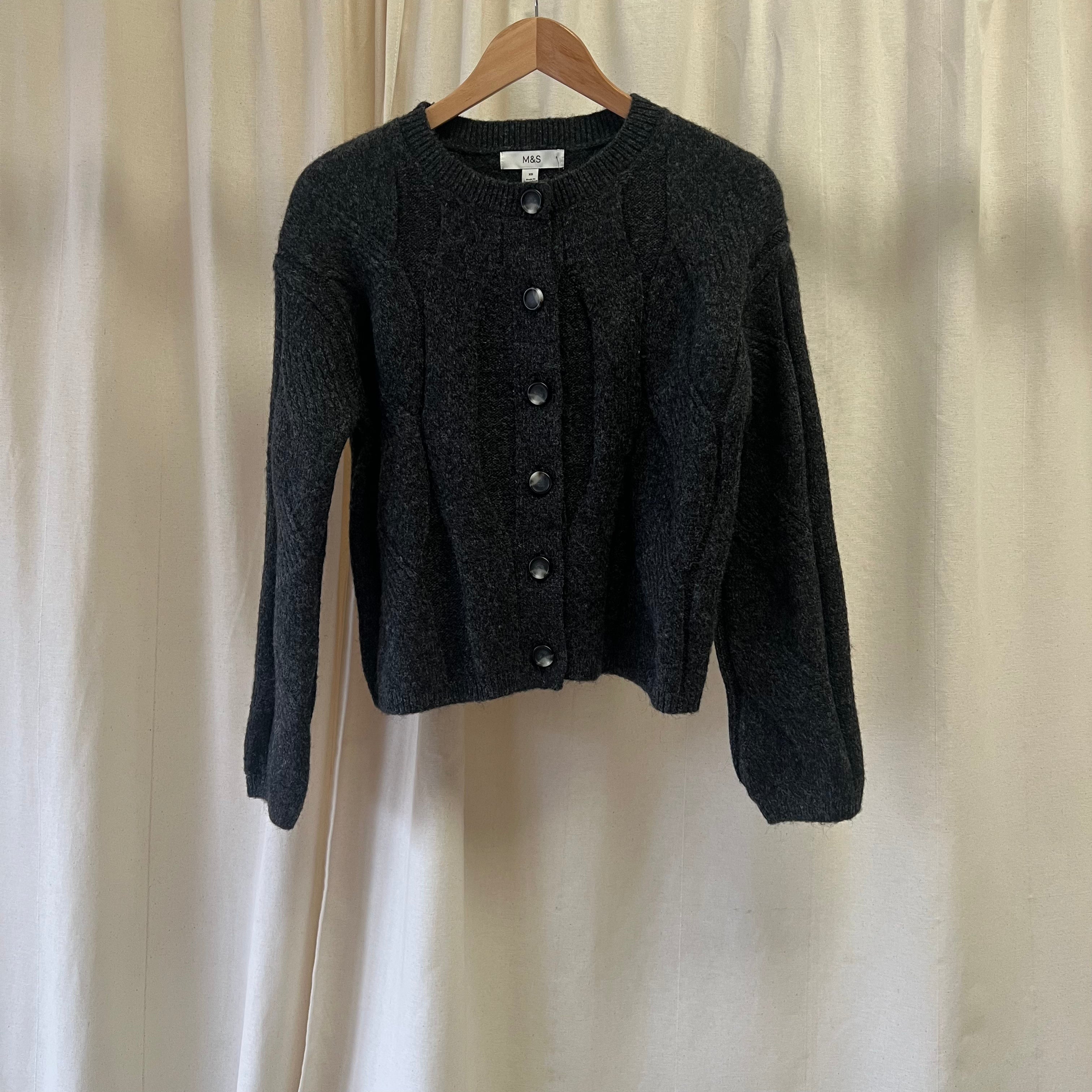 Dark gray cardigan on a wooden hanger against a beige curtain background