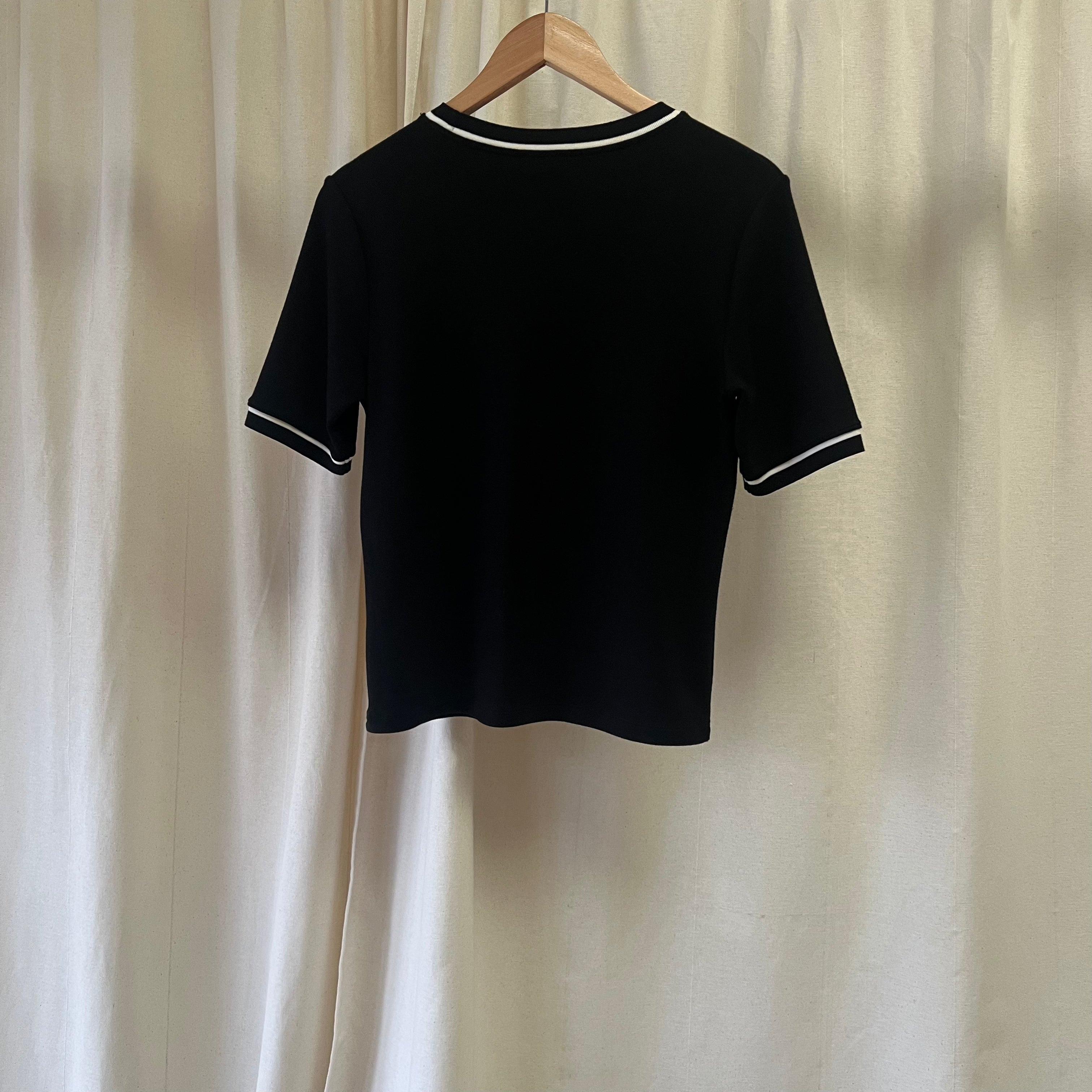 Black t-shirt with white trim on a hanger against a beige curtain background