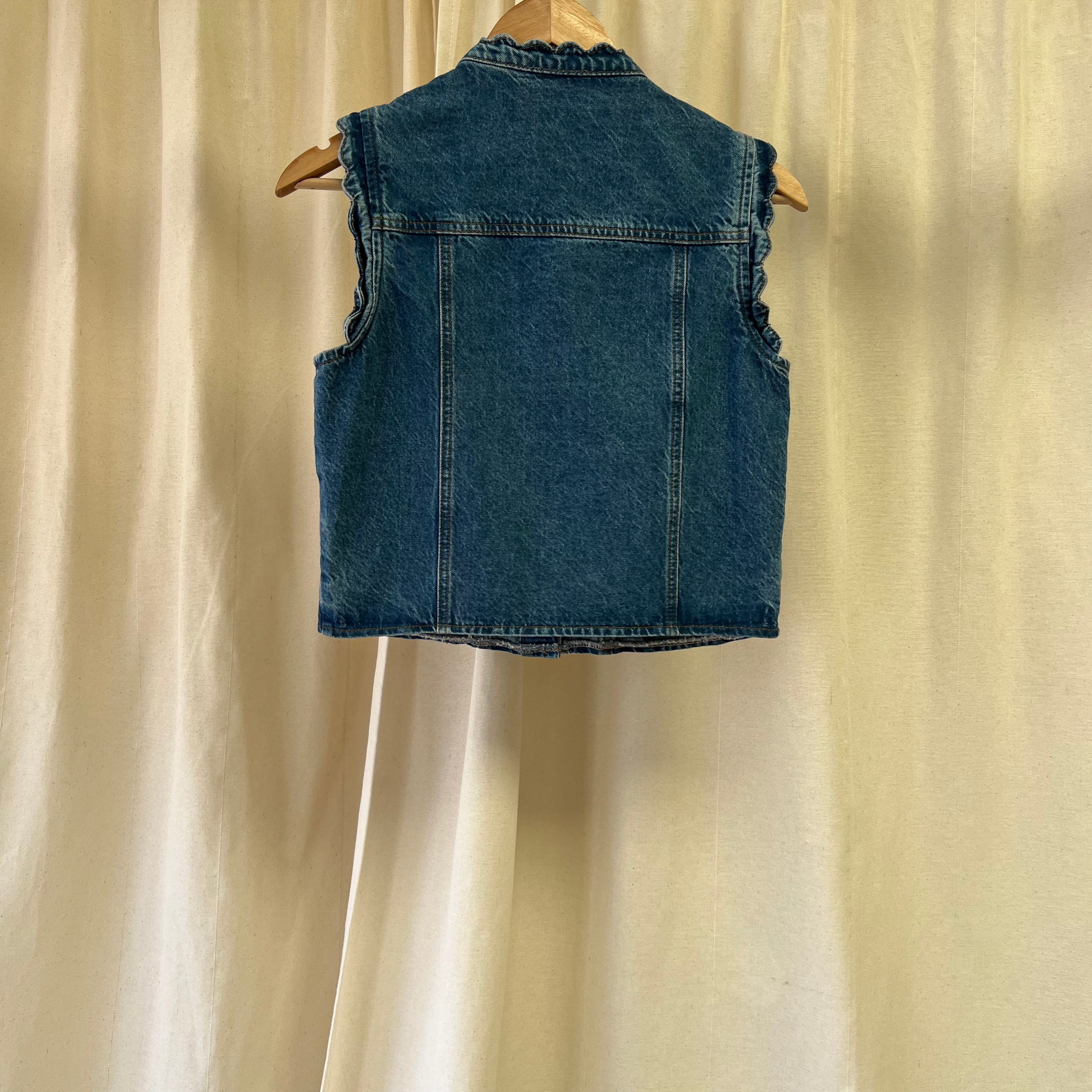 Blue denim vest on a wooden hanger against a beige curtain background
