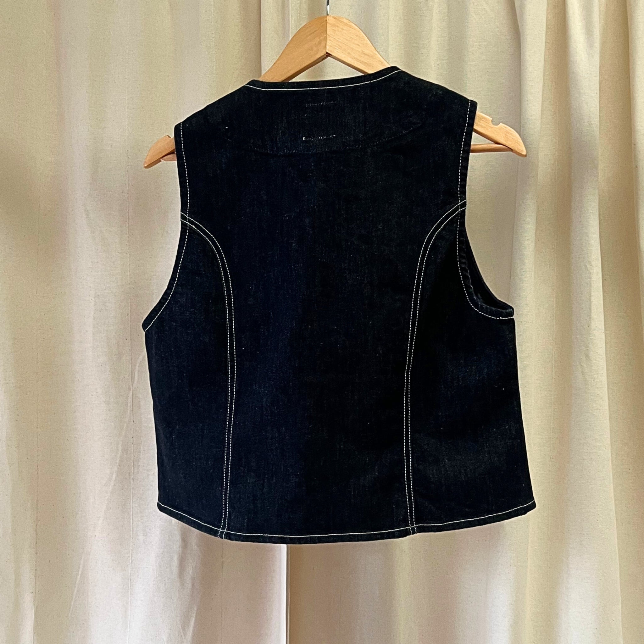 Black sleeveless vest on a wooden hanger against a beige curtain background