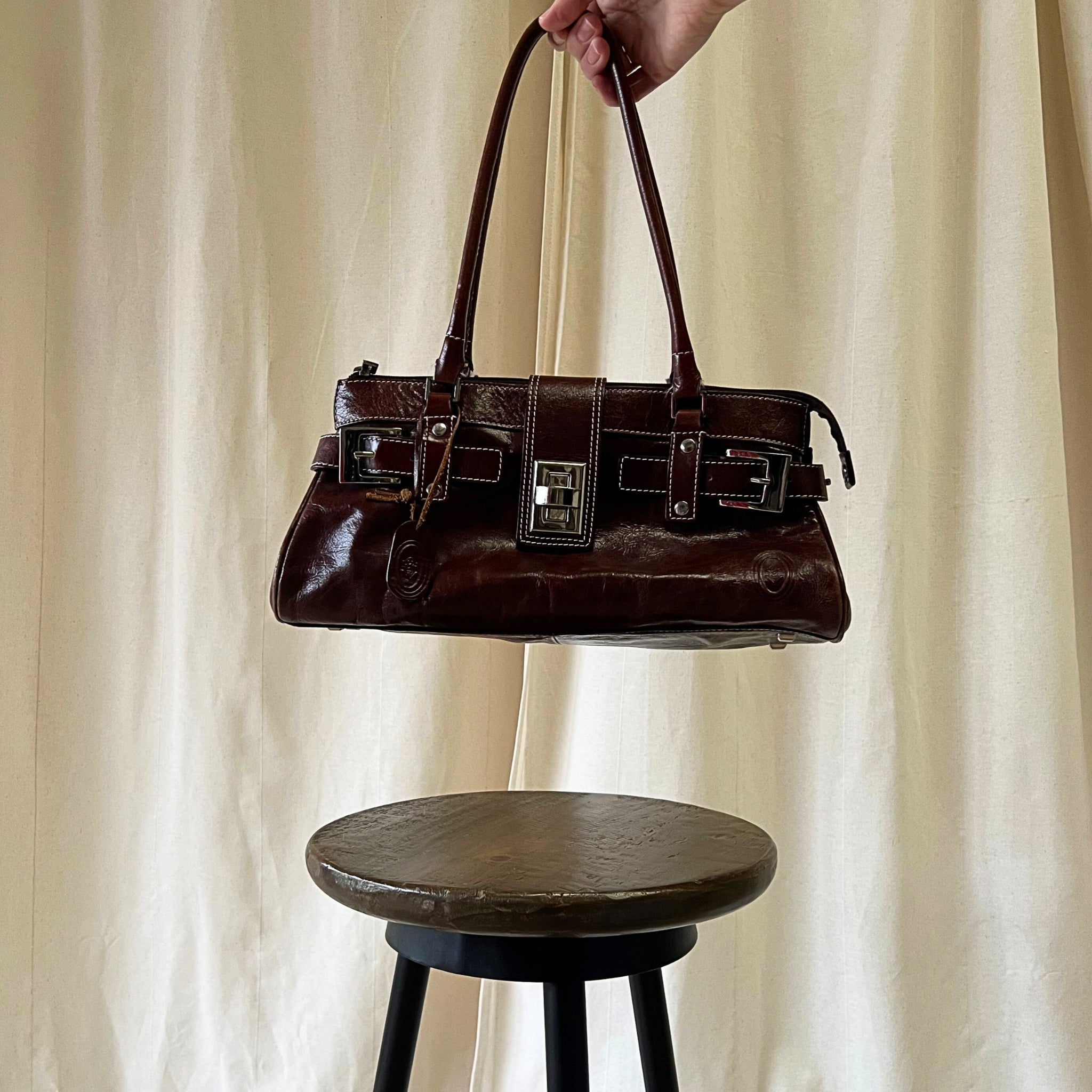 Brown leather handbag held above a wooden stool against a beige curtain background