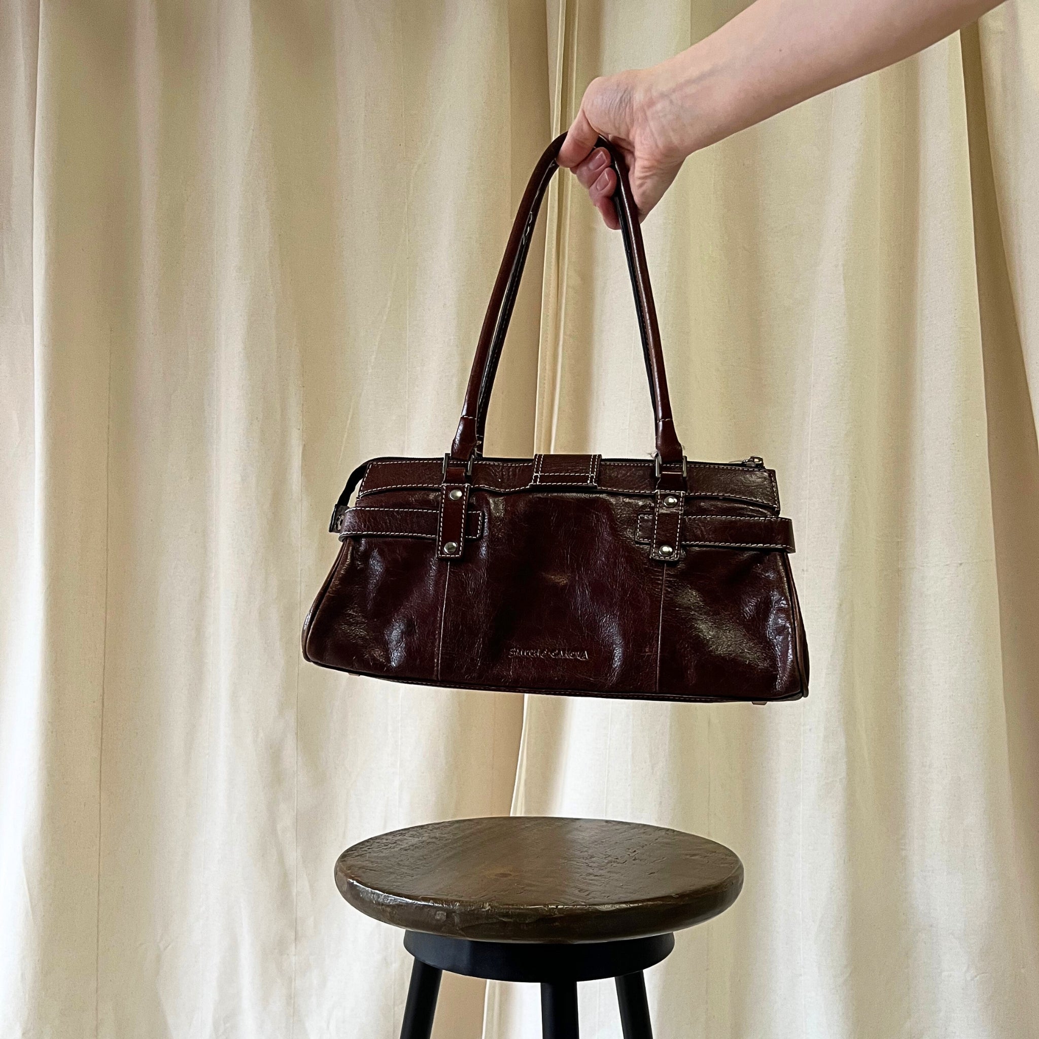 Brown leather handbag held above a wooden stool against a beige curtain background
