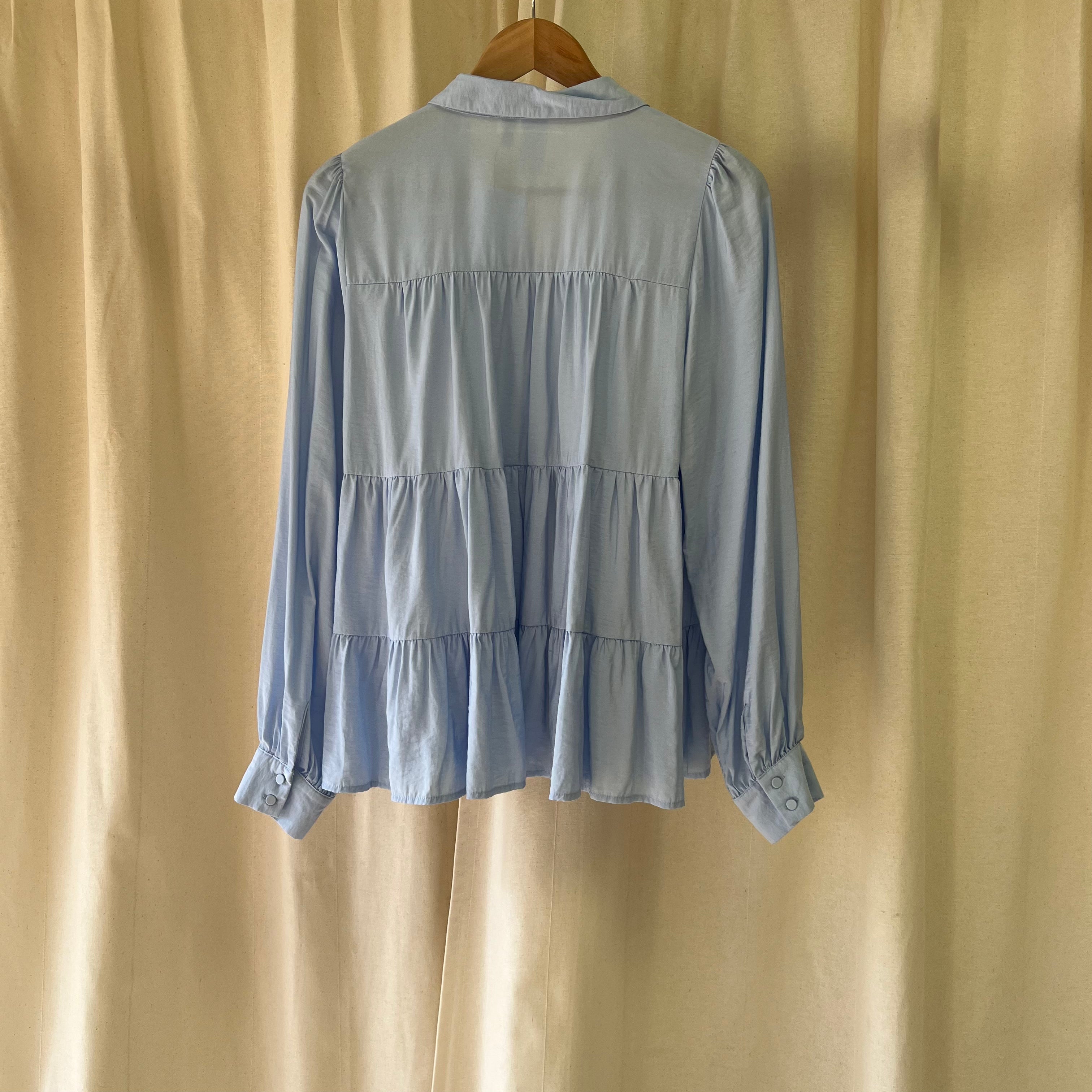 Light blue tiered blouse on a wooden hanger against a beige curtain background
