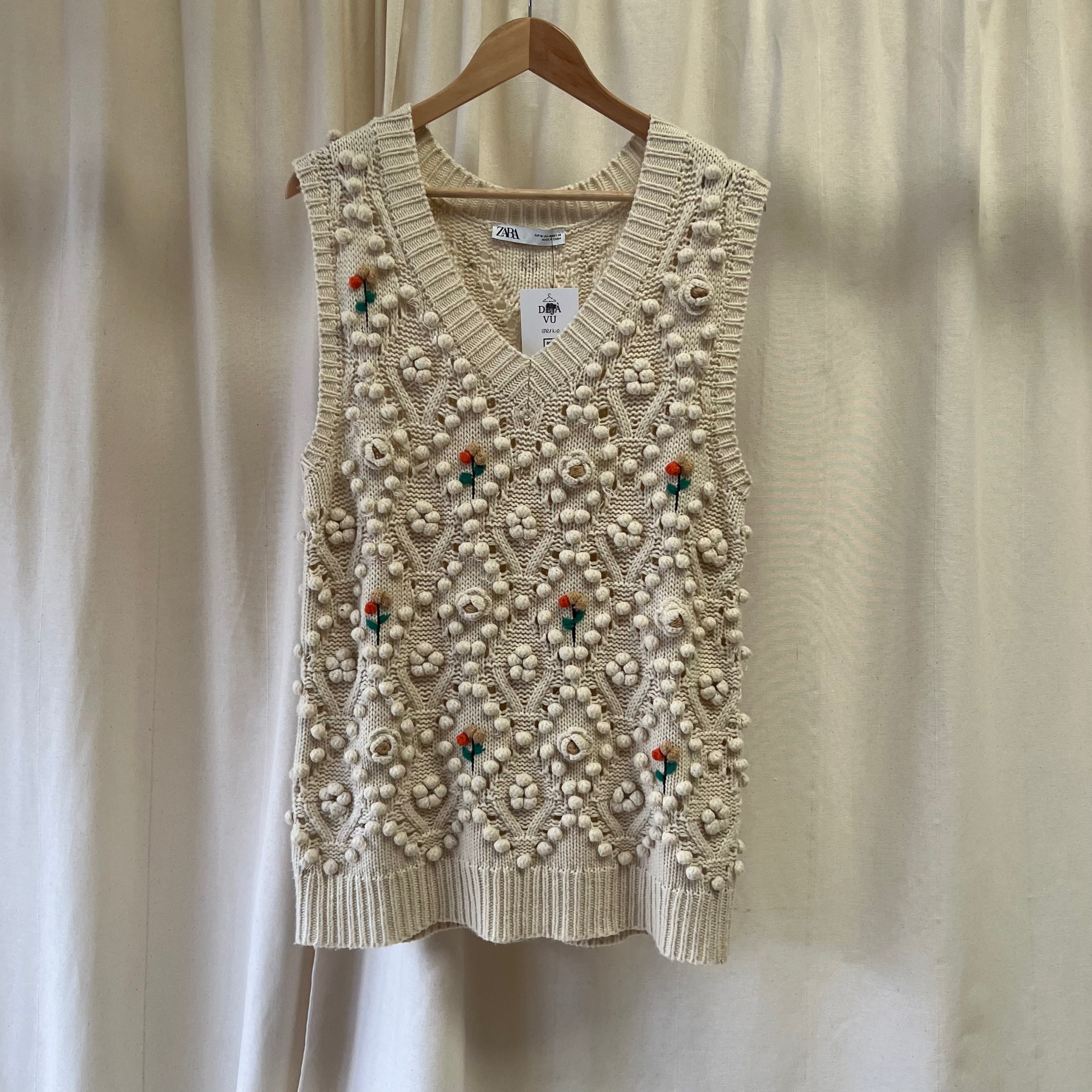 Beige knitted vest with floral patterns on a wooden hanger against a neutral background